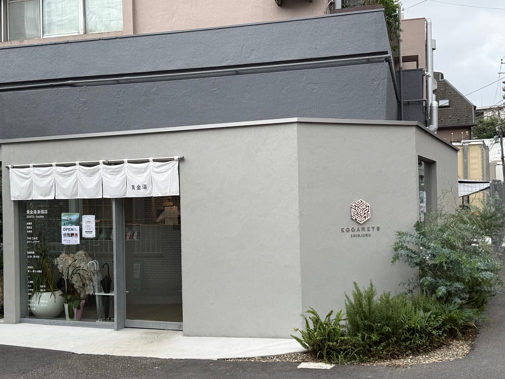 黄金湯 新宿店の入口付近の外観。白い暖簾と「KOGANEYU SHINJUKU」のロゴが見える。