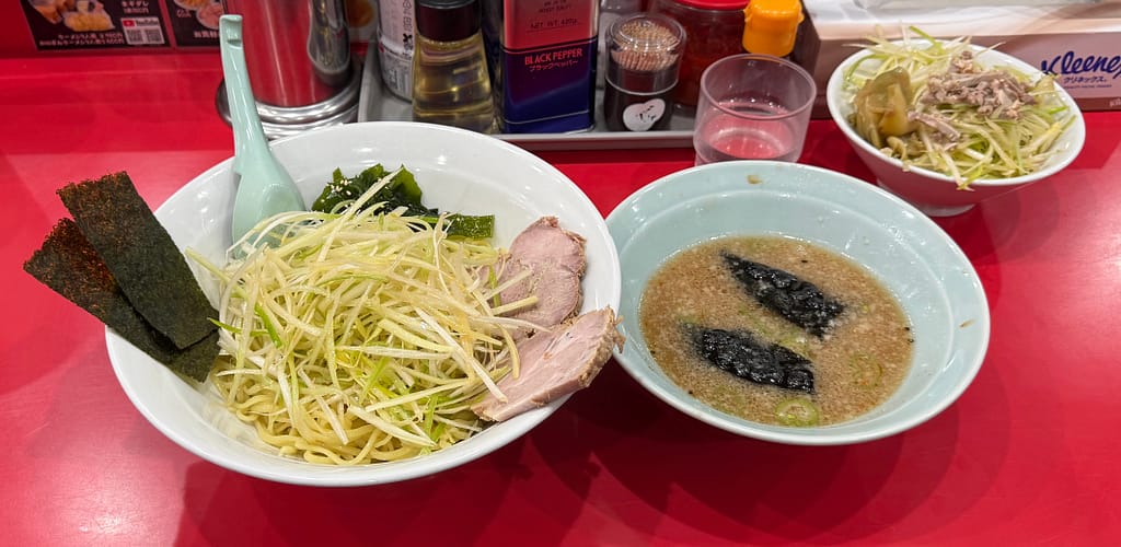 ラーメンかいざん 船橋店の「かいざんつけ麺」とネギチャーシュー丼。山盛りの白髪ネギが乗ったつけ麺と、別皿の濃厚つけ汁を赤いカウンター席で撮影した一枚。