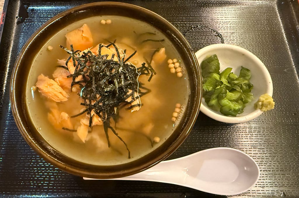 カプセルホテル&サウナ北欧のお茶漬け(鮭)。鮭、刻み海苔、あられがのったお茶漬けに、漬物とわさびが添えられた一品。