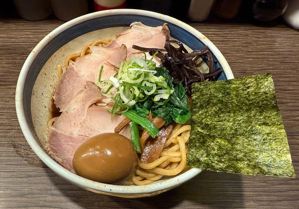 「油カダブラの特製油そば。チャーシュー、味玉、海苔、ほうれん草、ネギ、きくらげ、メンマが盛り付けられた豪華な油そばのアップ写真。」