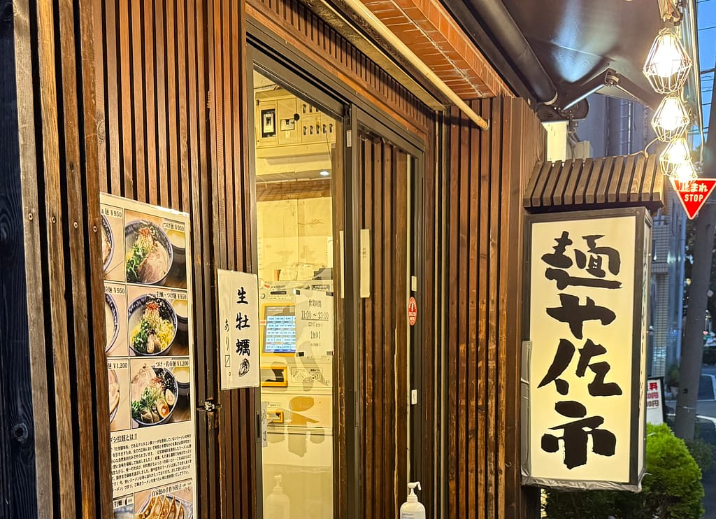 錦糸町の人気店「麺や佐市」の店舗外観。木目調の外壁と暖色照明、牡蠣ラーメンのメニューが掲示された温かみある入口