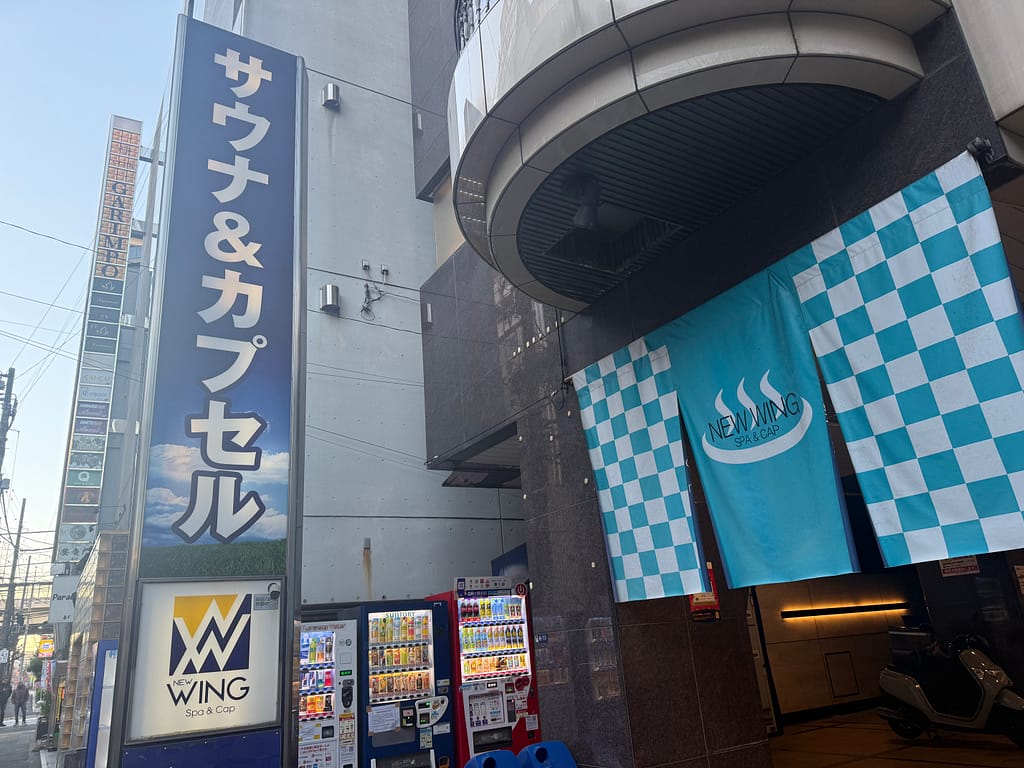 「『サウナ&カプセル』と大きく書かれた縦看板と、チェック柄の青い暖簾が掛かるニューウイング(NEW WING)入口。自販機が並ぶ建物前の様子を写した外観写真。」