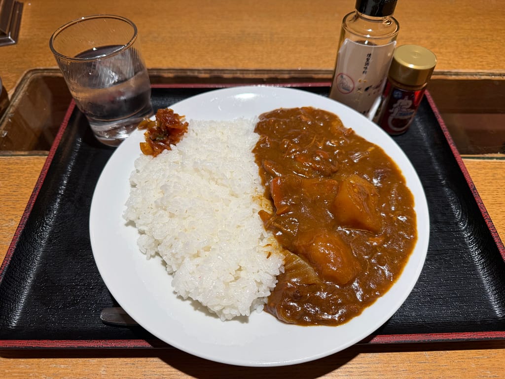 サウナ&カプセルホテル北欧の北欧特製カレーライス。トレーの上に白ごはんと具だくさんのカレーが盛られ、卓上調味料と水とともに提供されている。