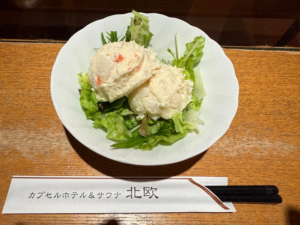 サウナ&カプセルホテル北欧のポテトサラダ。木目のカウンターに置かれた白い皿に、レタスと2玉のポテトサラダがきれいに盛り付けられている。