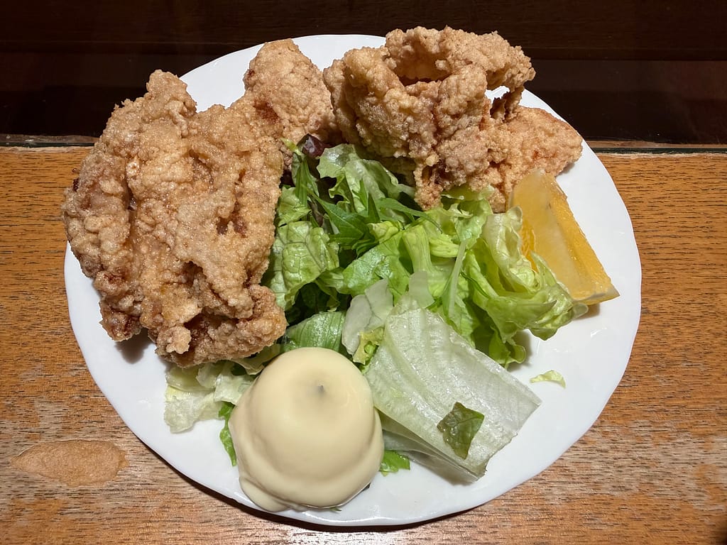 サウナ&カプセルホテル北欧の若鶏の唐揚げ。木目のカウンターに置かれた白い皿に、大ぶりの唐揚げとサラダ、マヨネーズ、レモンが盛り付けられている。