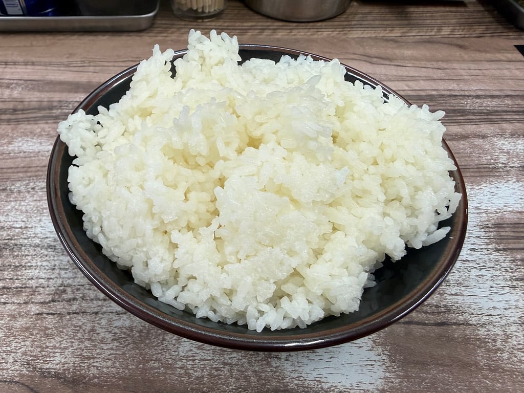 黒いラーメン丼に山盛りに盛られた白ごはん。粒立ちの良いツヤのある米がこんもりと盛られ、ボリューム感のある約700gのライスメガ盛り。木目調のカウンターの上に置かれ、迫力のある一杯。