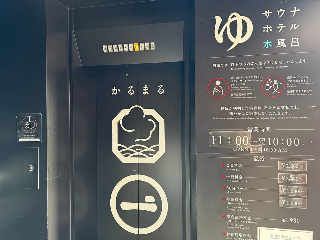 サウナ&ホテルかるまる池袋のエレベーター入口。黒を基調とした扉に「かるまる」のロゴと水風呂・サウナを示すアイコンが描かれ、右側には営業時間や利用案内が掲示されている