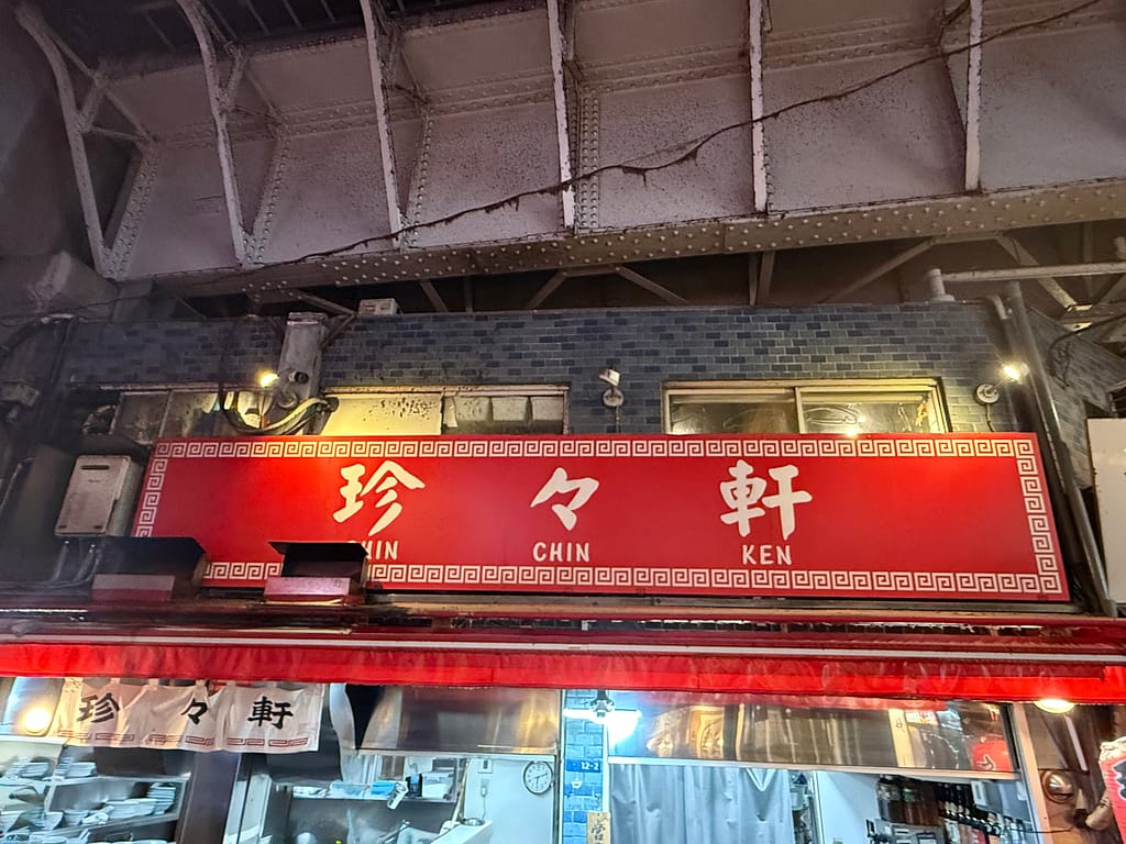 珍々軒の店舗外観。高架下に掲げられた赤い看板に「珍々軒(CHIN CHIN KEN)」の文字が映える老舗町中華。