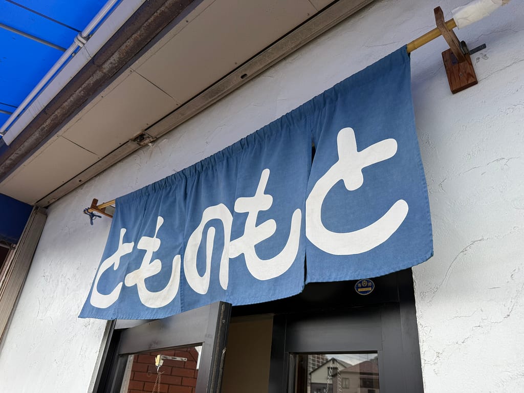 とものもとの店舗入口。青い暖簾に白文字で「とものもと」と書かれている。
