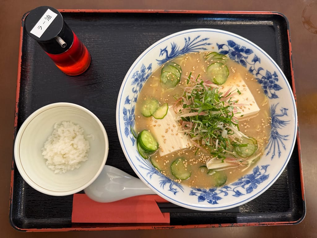 「冷や汁ラーメンのセット。白いご飯の小鉢と、赤い調味料ボトルが添えられ、きゅうり・豆腐・みょうが・ごまがのった冷たいスープのラーメンが黒いお盆に乗っている。」