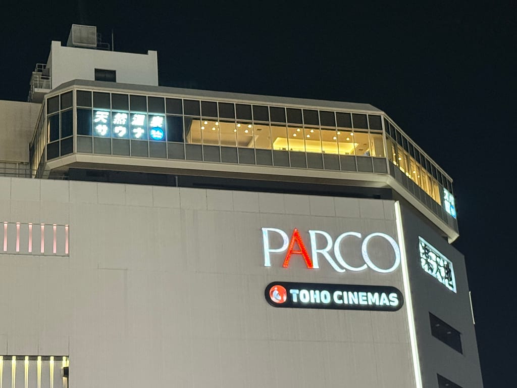 「錦糸町パルコの最上階にある『天然温泉 楽天地スパ』の夜の外観。ガラス張りの外観から明るい館内の光が漏れ、PARCOとTOHO CINEMASのロゴがビル壁面に見える。」