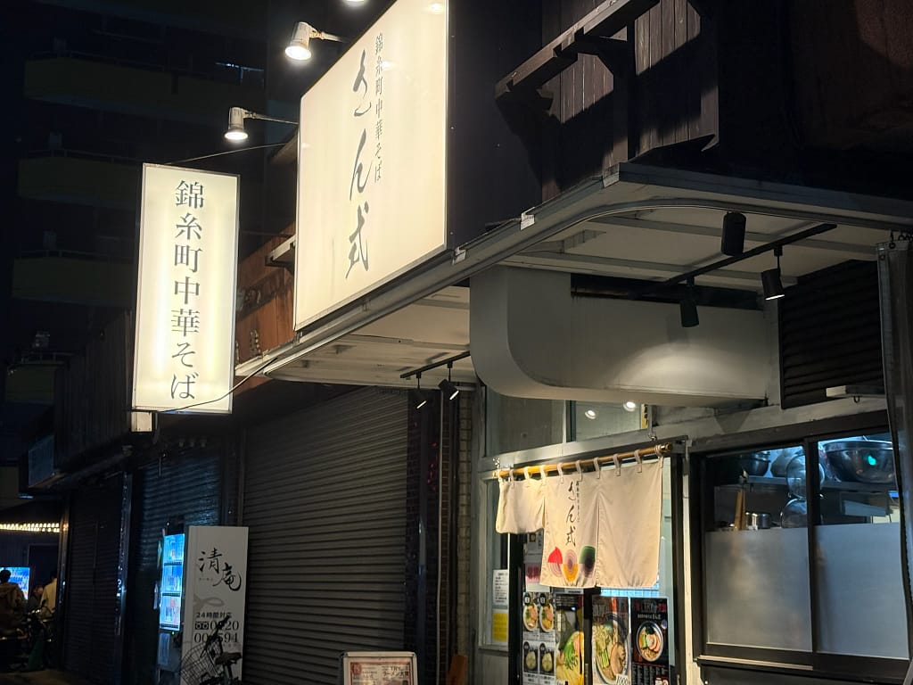 「夜の街に佇むラーメン店の外観。『錦糸町中華そば』と書かれた縦看板と、白地に屋号が書かれた大きな看板が並ぶ。入口には暖簾がかかり、店内の明かりが柔らかく漏れている。」