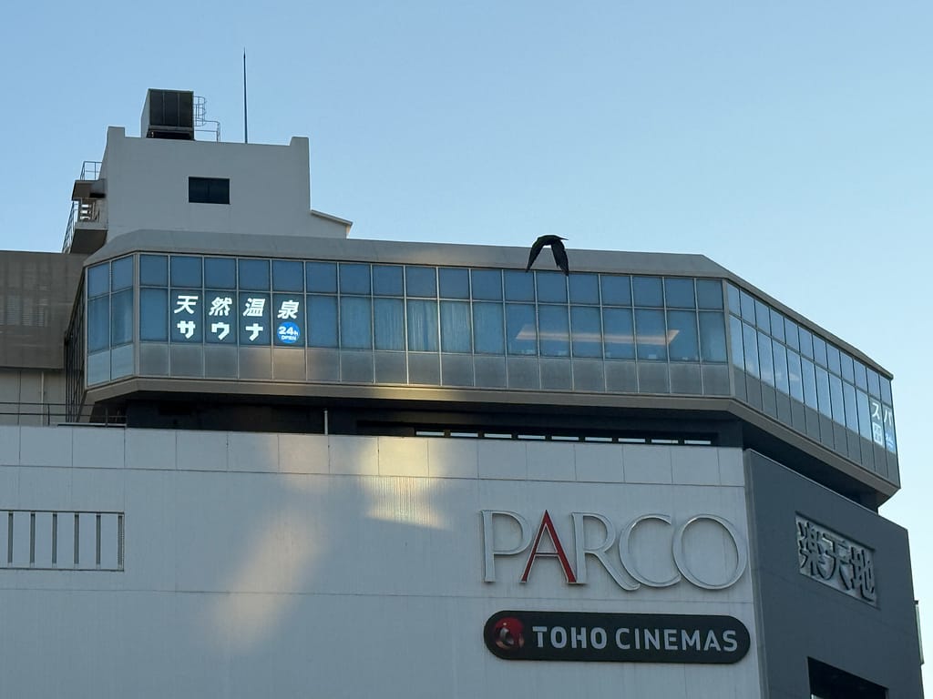 「ビル屋上に張り出すように設けられたガラス張りのサウナ施設。窓には“天然温泉・サウナ”と白文字で表示され、下層にはPARCOとTOHO CINEMASの看板が見える朝方の外観。」