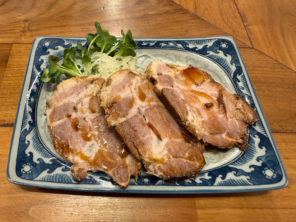 麺や佐市のつまみ豚肉。しっとり柔らかく仕上げた厚切りチャーシューにタレがかかり、カイワレとネギが添えられた一品。