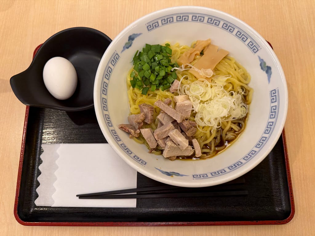 レインボー新小岩のレストランで提供されているカレーまぜそば。醤油ベースのタレが絡んだ麺に、角切りチャーシュー、白ネギ、青ネギ、メンマが彩りよく盛り付けられている。別皿には生卵が添えられ、シンプルながらサウナ後に食欲をそそる一杯。