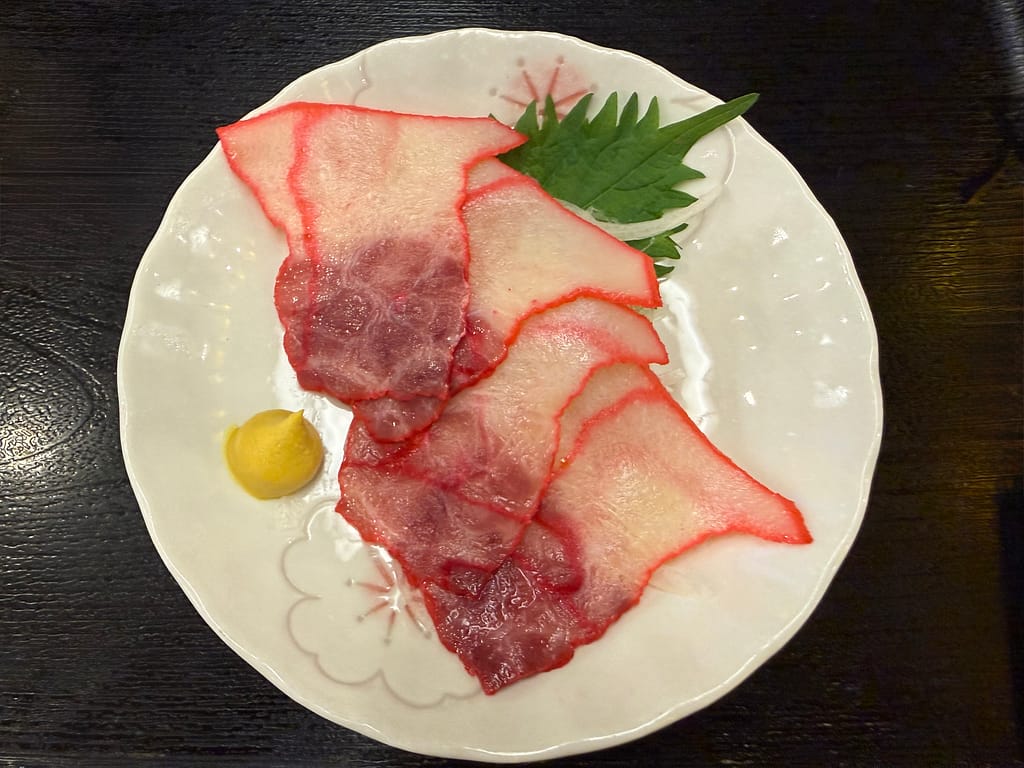 レインボー新小岩のレストランで提供されているクジラベーコン。赤い縁取りが特徴的な薄切りのクジラベーコンが美しく盛り付けられ、からしと大葉が添えられている。サウナ後の一品料理として、昔ながらの居酒屋の雰囲気を感じさせるメニュー。