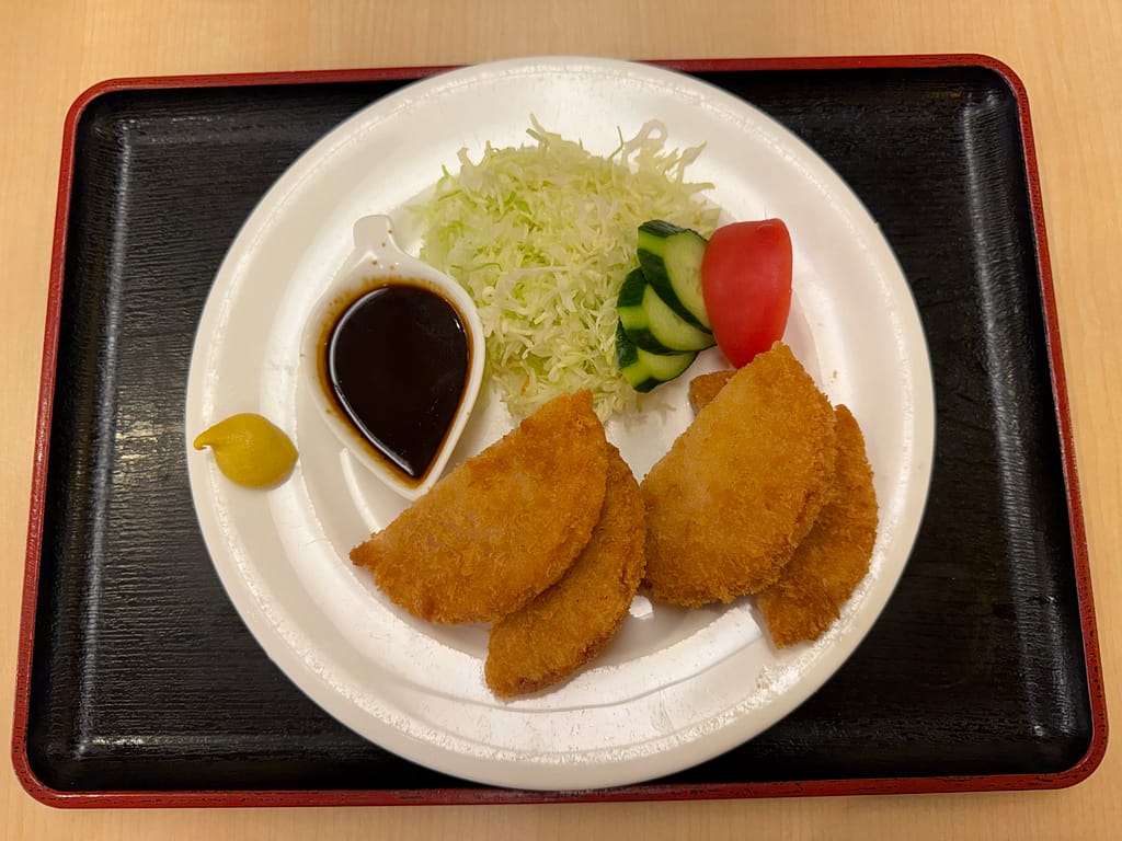 レインボー新小岩のレストランで提供されているハムカツ。衣はサクサクに揚げられており、添えられたソースとからしで味わえる一品。千切りキャベツ、きゅうり、トマトが添えられ、サウナ後の食事やおつまみにぴったりの昔ながらの定番メニュー。