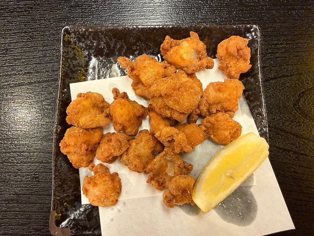 レインボー新小岩のレストランで提供されている軟骨唐揚げ。カリッと揚げられた一口サイズの軟骨唐揚げが黒い皿に盛り付けられ、添えられたレモンが爽やかなアクセントになっている。サウナ後のおつまみとして相性抜群の定番メニュー。