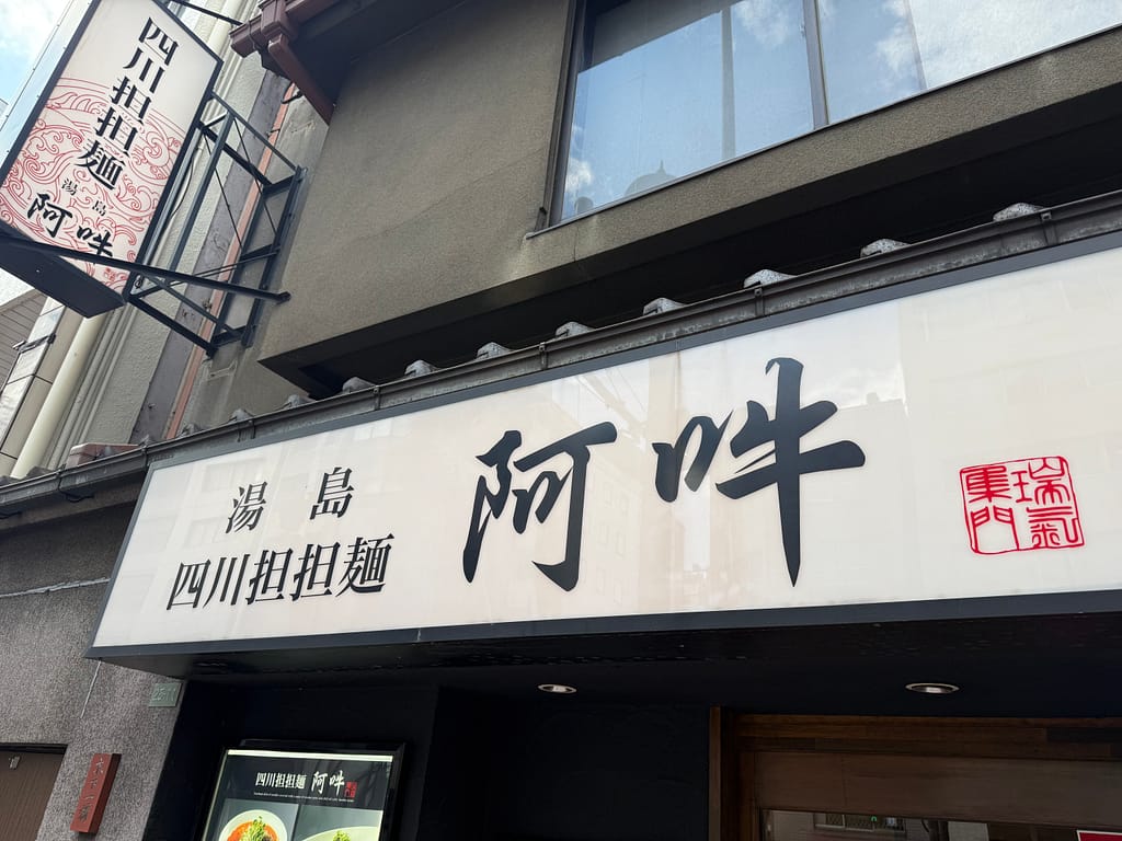 湯島の担担麺専門店「四川担担麺 阿吽」の店頭看板。白い看板に大きく「阿吽」と黒い筆文字で書かれ、横に赤い印章風のマークがある。シンプルで落ち着いた外観。