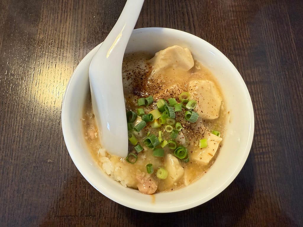 小さな白い器に盛られたマーボー丼（小）。白い豆腐がとろみのある餡に包まれ、上には刻みネギと花椒がふりかけられている。スープのように優しい見た目ながら、香り立つ痺れが食欲をそそる一品。