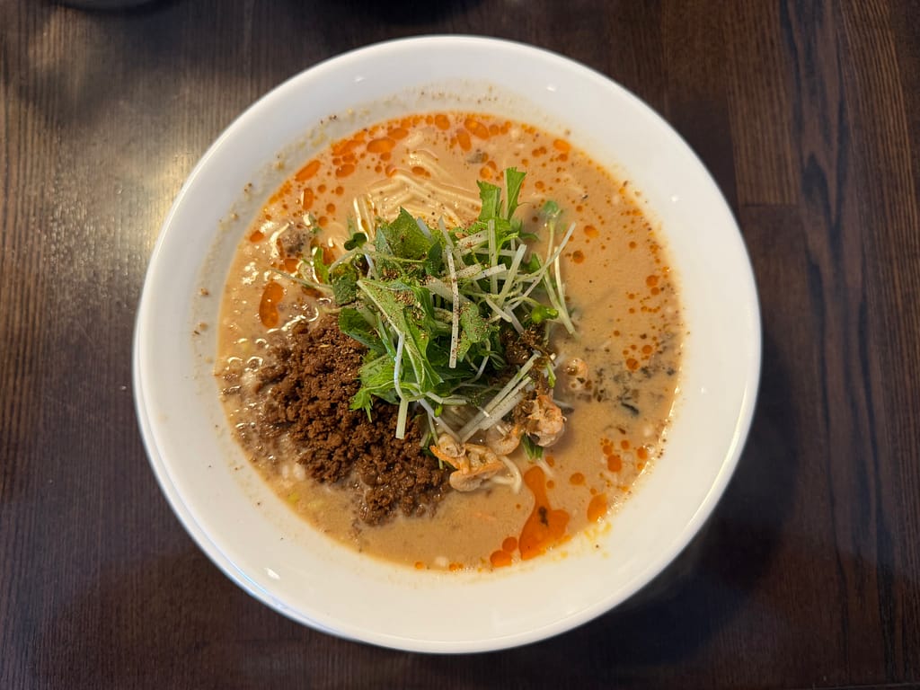 白い器に盛られた「阿吽」の白胡麻担々麺。表面には鮮やかな辣油が広がり、中央には肉味噌、水菜、干しエビなどの具材が彩りよく盛り付けられている。クリーミーな胡麻スープに香ばしい香りが立ちのぼる一杯。