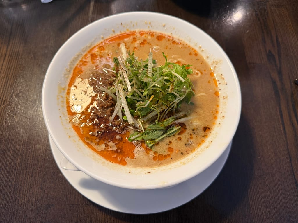 白い丼に盛られた白胡麻ベースの担担麺。濃厚な胡麻スープに赤いラー油が浮かび、肉味噌と刻んだ青菜・細切りネギが中央に乗っている。辛さ2・痺れ2で、まろやかさの中にスパイスが香る見た目。