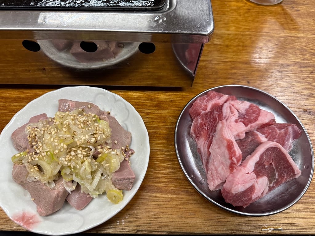 マトンとレバーの盛り合わせ。左の皿には白ごまを散らしたレバーが山盛りに盛られ、右の皿には鮮やかな赤身のマトン肉が並んでいる。焼き台の前に置かれた調理前の食材が写っている。