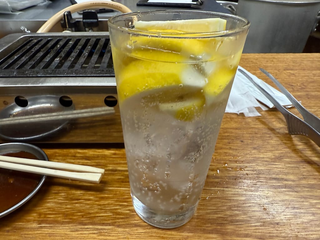 「グリルのあるカウンター席に置かれたレモンサワーのグラス。中には輪切りのレモンがたっぷり入っており、炭酸の気泡が立ち上る様子が見える。」