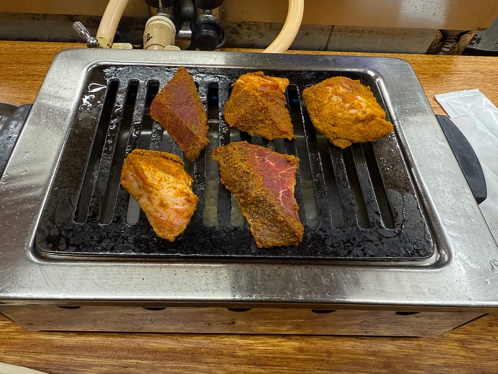 「卓上の小型グリルで焼かれているスパイスたっぷりのマトン肉。表面がスパイスで色づいた大きめの肉片が数個並び、焼き目がつき始めている様子。」