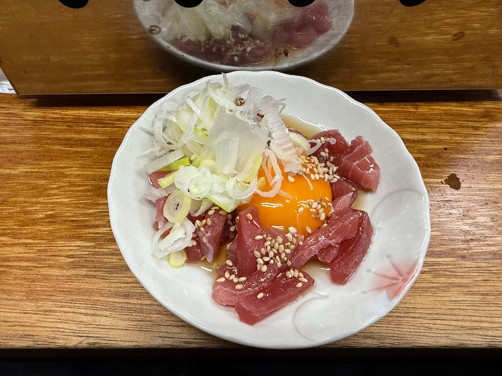 「白い小皿に盛られたマグロユッケ。角切りのマグロの赤身に卵黄、刻みネギ、白ごまが乗り、木目のカウンターの上に置かれている。」