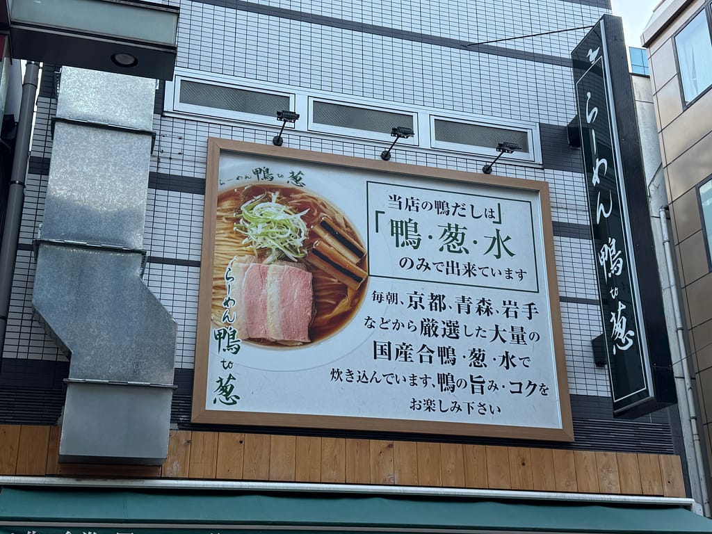 鴨と葱、水のみで出汁を取ることを掲げたラーメン店「らーめん 鴨 to 葱」の外観看板。鴨だしラーメンの写真とこだわりが記された大きな看板。