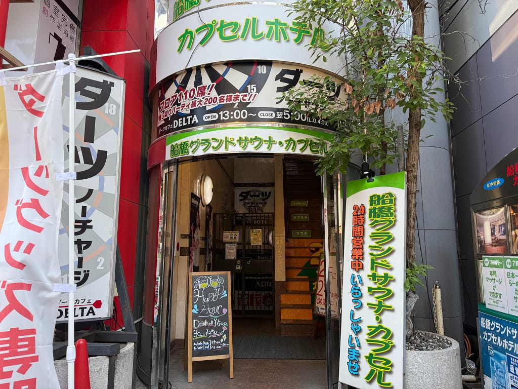 船橋グランドサウナの入口外観。カプセルホテルの看板とラーメンを掲げたサウナ施設の入口。