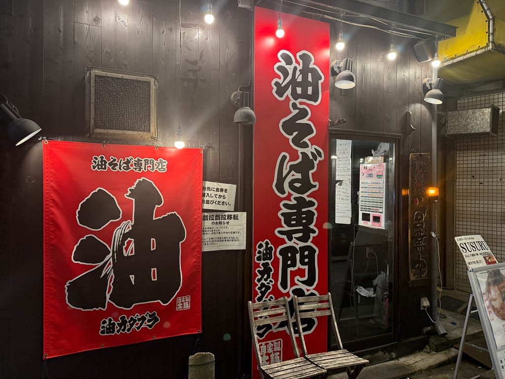 「船橋の油そば専門店『油カダブラ』の外観。赤い大きなのぼりと暖簾に“油そば専門店”の文字が入り、夜のライトに照らされた店前の様子。」