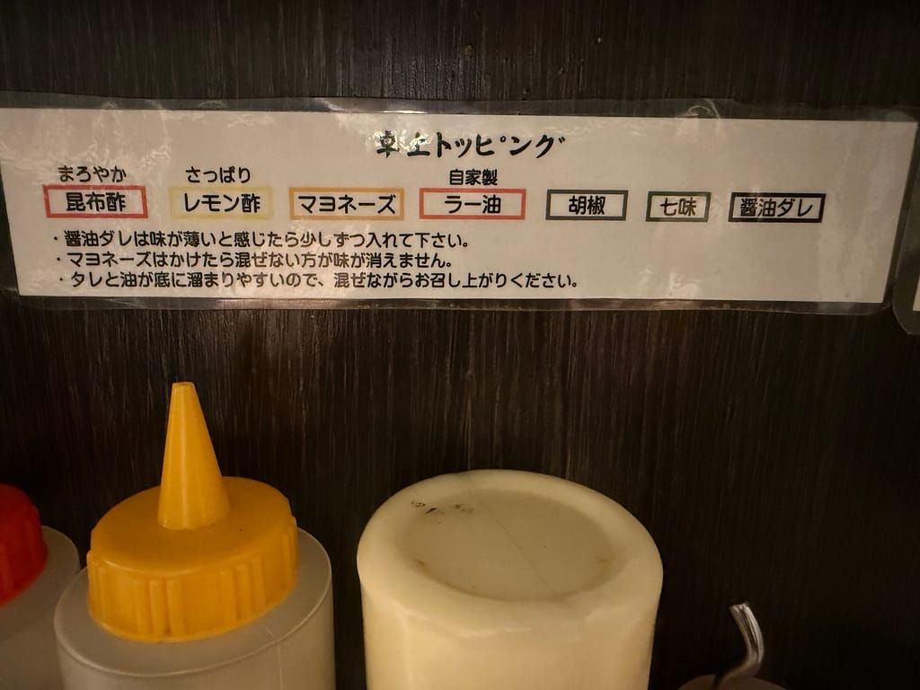 「油カダブラの卓上トッピング説明。昆布酢、レモン酢、マヨネーズ、自家製ラー油、胡椒、七味、醤油ダレの使い方を案内する張り紙と、下に並ぶ調味料ボトル。」