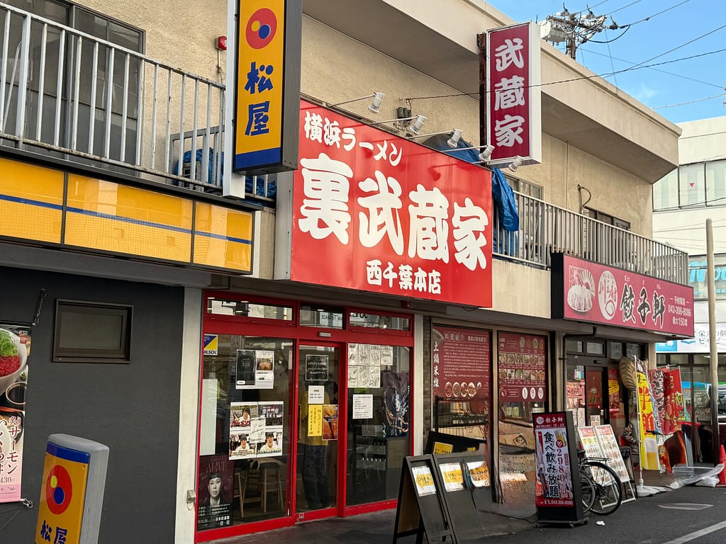 西千葉駅南口から徒歩1分にある「裏武蔵家 西千葉本店」の外観。赤い看板に白文字で「裏武蔵家」と書かれ、横浜家系ラーメンの文字が目立つ店舗正面の様子。
