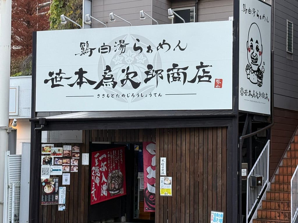 鶏白湯らぁめん 笹本為次郎商店の外観。白い看板に筆文字で店名が書かれ、入口横には店主のイラスト看板が掲げられている。