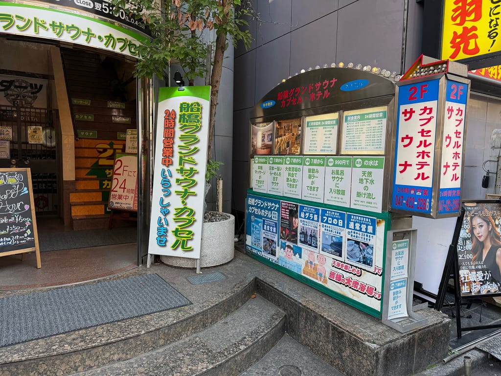 「船橋グランドサウナ入口。『日本一旨いらーめんを出すサウナを目指しています』と書かれた掲示板と、料金・サービス案内が写っている外観写真。」