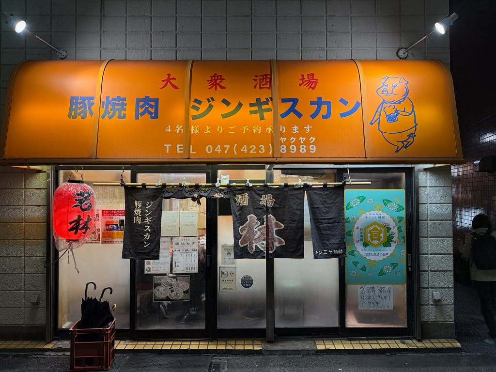 船橋の大衆酒場「豚焼肉 ジンギスカン」の夜の外観、オレンジ色の看板と暖簾が目印の居酒屋