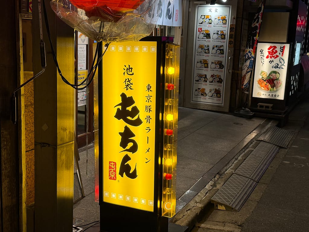 「池袋・屯ちん池袋本店の黄色い電飾看板。『池袋 東京豚骨ラーメン 屯ちん』と書かれた縦長の看板が夜の路地に光っている」