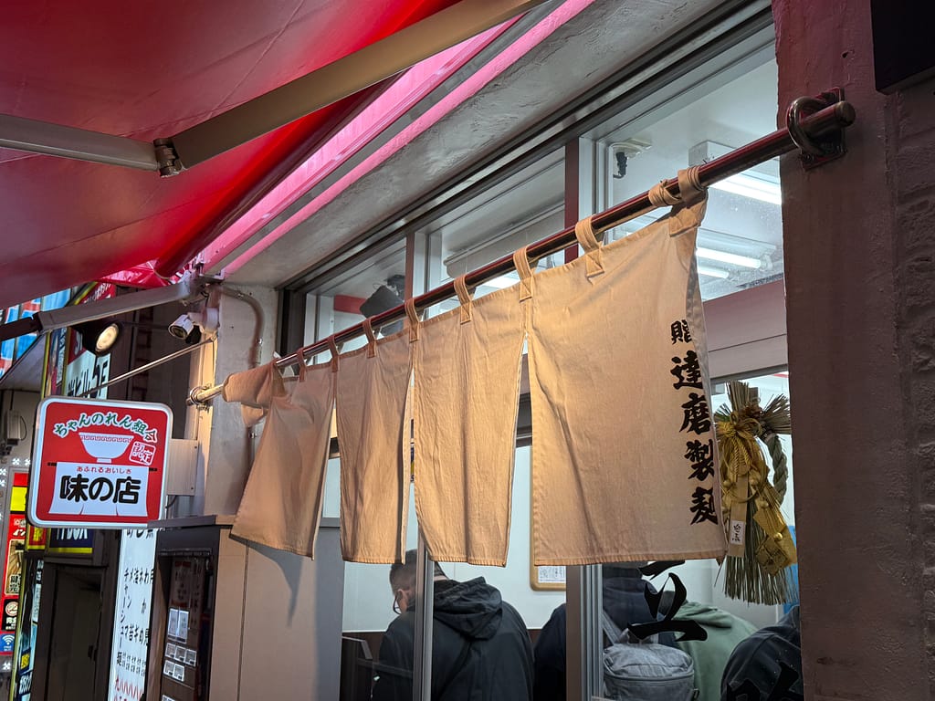 新橋ニューともちんラーメン蒲田西口駅前店の入口のれん。立ち食いスタイルの店内で客がラーメンを食べている様子が見える外観。