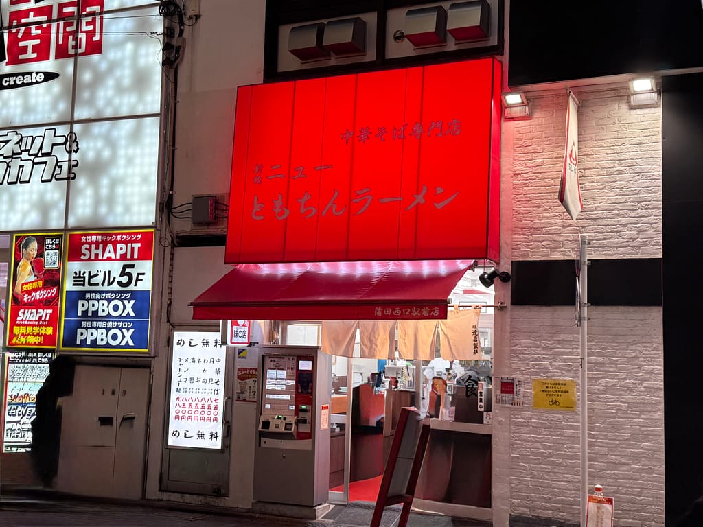 新橋ニューともちんラーメン蒲田西口駅前店の外観。赤い大きな看板が目印の立ち食いラーメン店で、入口の券売機と店内の様子が見える夜の店舗風景。