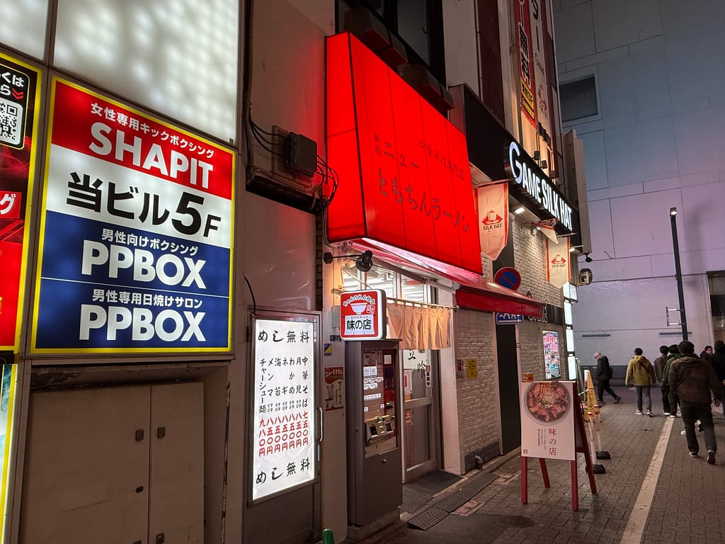 新橋ニューともちんラーメン蒲田西口駅前店の外観。赤い看板が目印の立ち食いラーメン店で、夜の蒲田の街並みに面した店舗入口と券売機の様子。