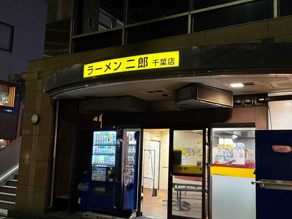 ラーメン二郎千葉店の外観。夜の時間帯に黄色い看板が光り、入口横には自動販売機が設置されている。