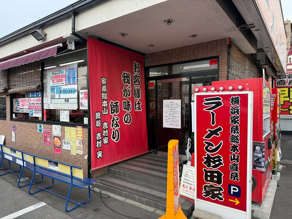 「杉田家 千葉祐光店の入口外観。赤い暖簾と看板が目印の横浜家系ラーメン直系店で、行列用ベンチや券売機の案内が見える」