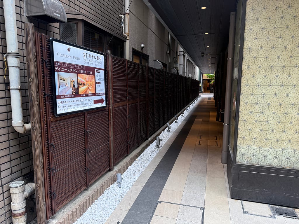 センチュリオンホテル&スパ上野駅前 本館エレベーターへ続く通路、木目調の壁と案内看板がある細い通路