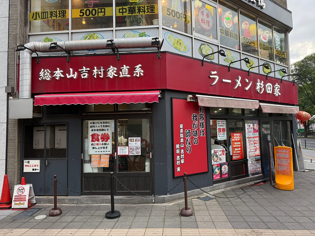 赤を基調とした「ラーメン杉田家千葉駅前店」の外観。店舗正面には「総本山吉村家直系」の看板が掲げられ、角地に位置するため視認性が高い。店前には行列整理用のポールが設置されている。