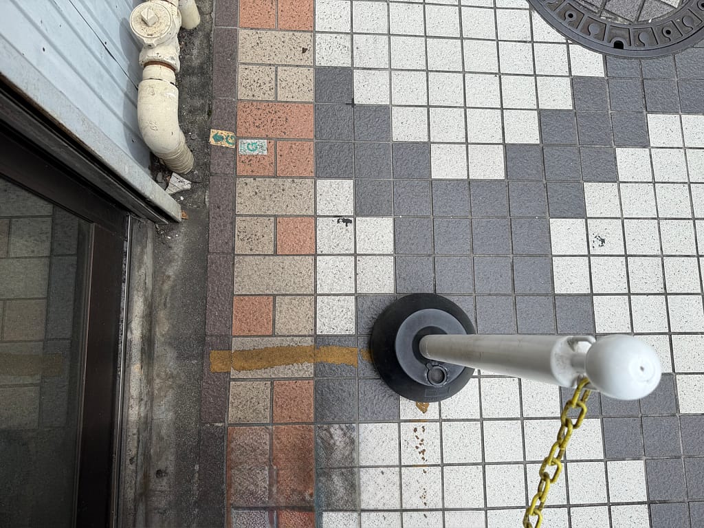 ラーメン二郎小岩店の行列の先頭位置を示す。タイル張りの地面に黄色い目印があり、チェーン付きのポールが設置されている。