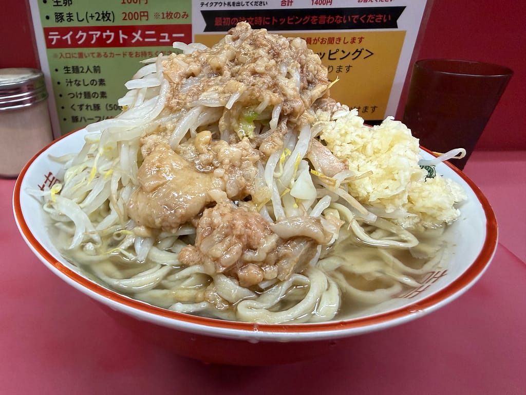 ラーメン二郎小岩店のラーメン。山盛りのもやしとキャベツの上に背脂がたっぷりとかかり、分厚い豚肉と大量の刻みニンニクが添えられている。太麺がスープから顔をのぞかせている。