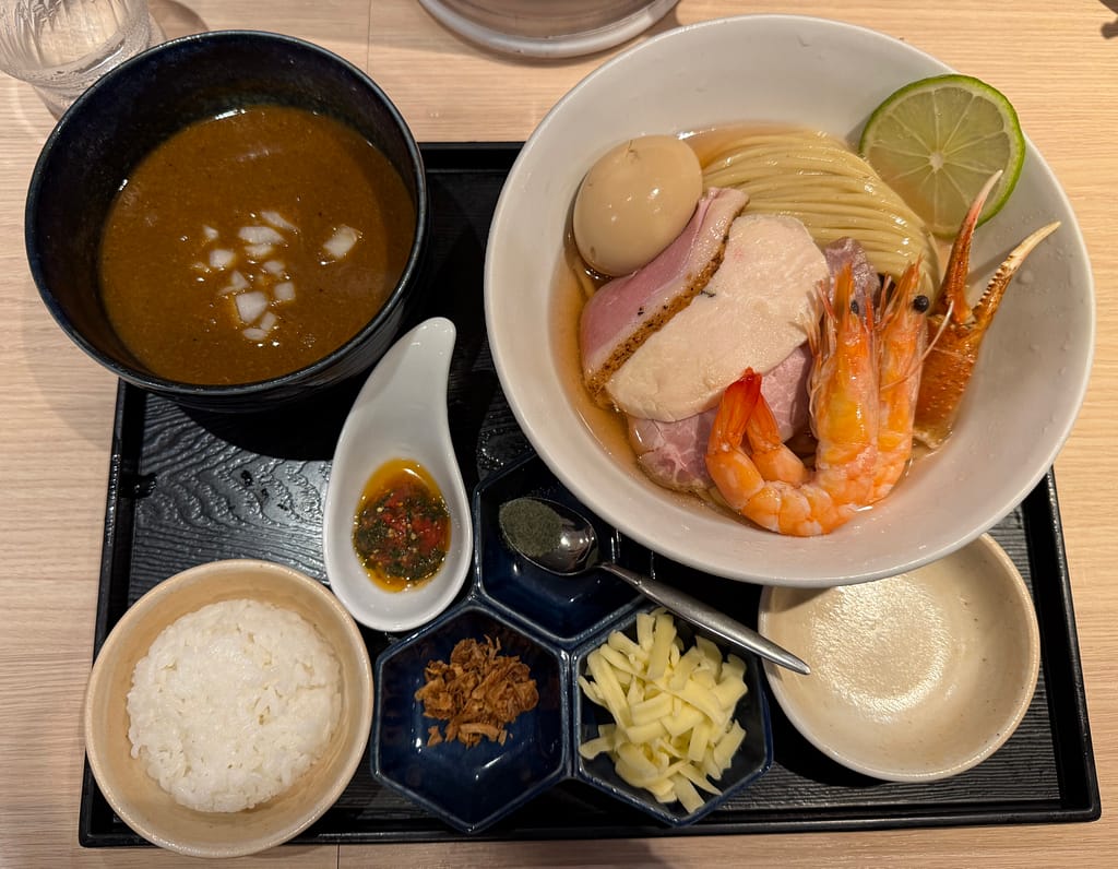 麺屋一燈の「オマール海老のビスクつけ麺 特製濃厚」。海老や3種のチャーシュー、煮卵、ライムが彩る美しい盛り付けに、濃厚なビスクスープとライス、チーズ、ソースピストゥなど多彩な付け合わせが並ぶ。