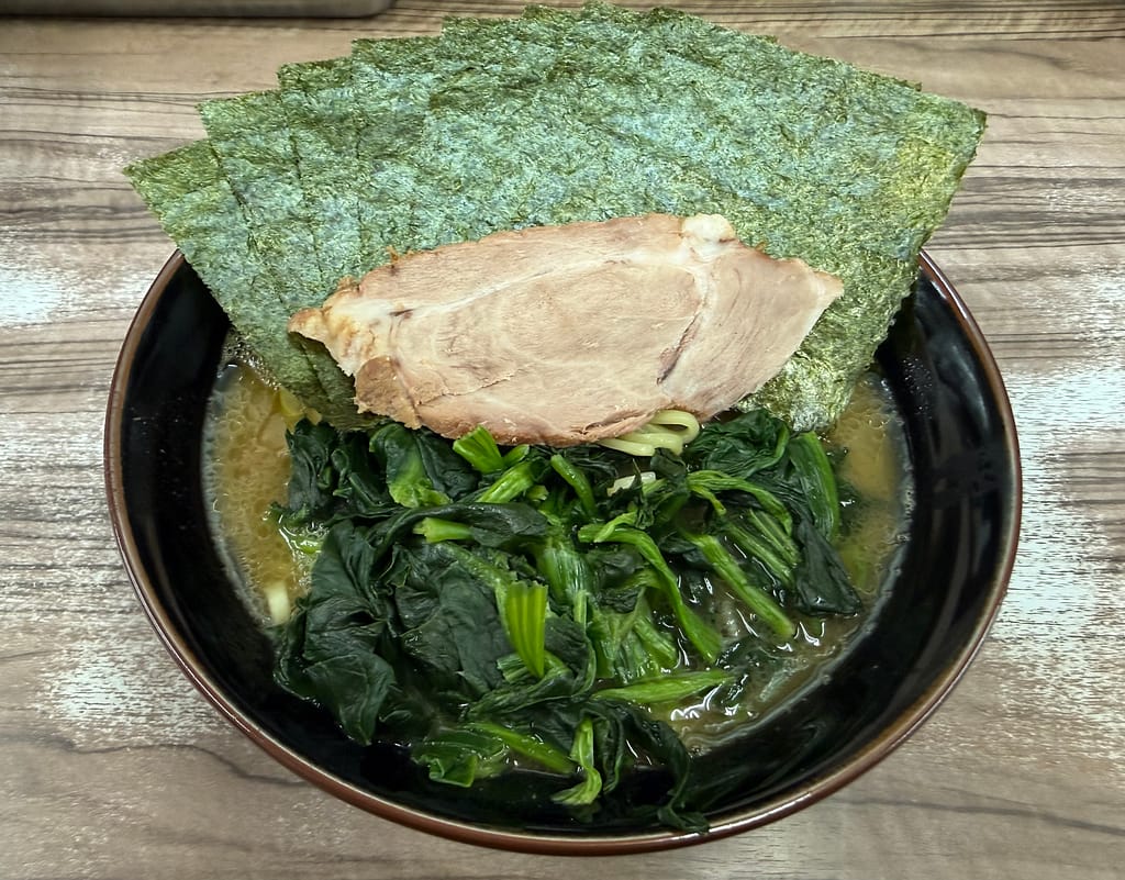 黒い丼に盛られた家系ラーメン。濃厚なスープの上にたっぷりのほうれん草が山のように乗り、その上に大判のチャーシューが一枚。さらに丼の縁を覆うように大きな海苔が複数枚トッピングされている。木目調のテーブルの上で、ボリューム感と迫力のある一杯。